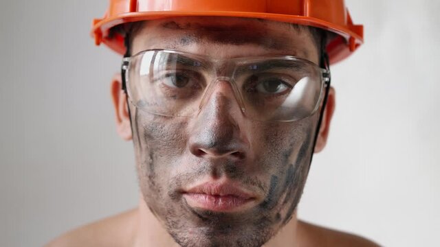 Portrait of a dirty tired male builder in a work helmet and glasses looks at the camera on a white background. Weary man construction worker.
