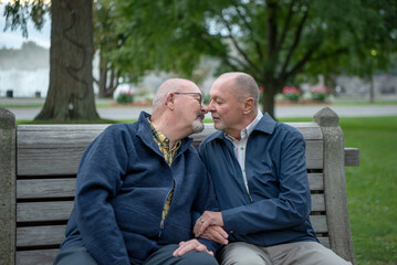 Elderly gay couple sitting on a park bench lean in to kiss each other.   One has his arms linked...