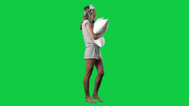 Side view of young woman in sleepwear with sleep mask holding pillow. Full body on green screen chroma key background