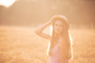 Portrait of a young, happy woman in a straw hat, pink long dress stands in a golden wheat field