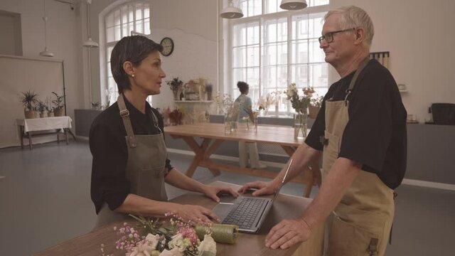 Medium shot pf two mature owners of small flower studio chatting while standing at counter. Female customer choosing items to buy in background