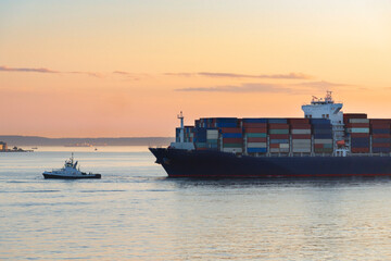 Tug boat bringing a shipping container ship into port at sunset. 