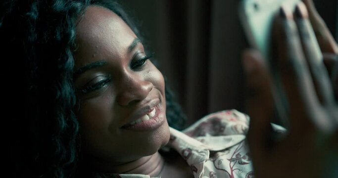 Super close-up portrait of a young attractive African American woman with curly hair lying on a sofa laughing after recieving a meassage while texting on the smartphone device in hands. Thining of a