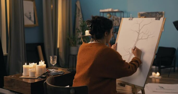 A woman with long brown hair pinned up in a bun using pencils sits by candles in the evening. A focused girl with the soul of an artist draws a picture of a tree, sketches on a cavas