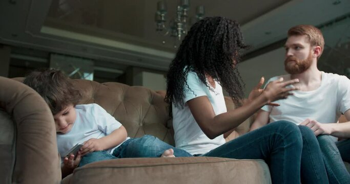 Sad little boy seated playing games on a phone whilst his parents fight at home in the living room.