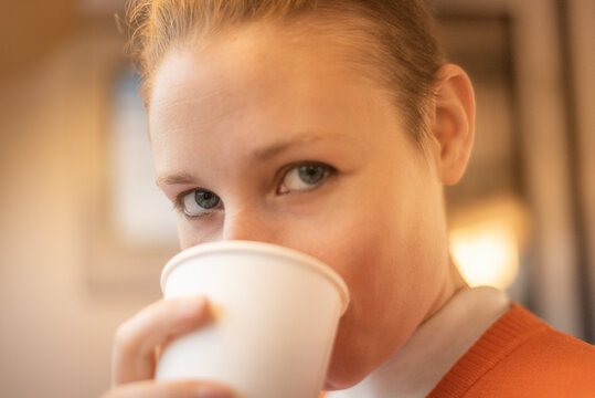 Young adult blonde caucasian woman with blue eyes, drinking from a takeaway paper cup looking at the camera. Selective focus in one eye