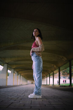 Full body portrait of Caucasian woman. The setting is urban with a symmetrical background with a walkway with pillars on the sides