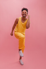 Happy muscular african guy rejoices in victory by clenching fists tightly on pink background. Model with dark curly hair wearing glasses wearing summer yellow suit and white shoes. Concept of emotions