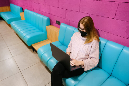 Young attractive woman working on laptop in modern coworking space with bright magenta walls and blue sofas and drinking cocoa. Elerning or freelance concept.