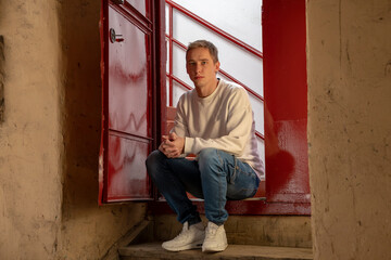 A young guy 20-25 years old with a backpack is sitting on the steps at the entrance to the entrance. Open entrance doors. Concept: fired from work, paid for housing, kicked out of the apartment.