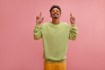 Hopeful african guy crossed his fingers and smiley praying for good exam results. Portrait shot of boy in stylish clothes in bright leafy and lemon colors, posing in front of isolated raspberry wall.