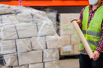 Close up of Warehouse Staff wearing a protective face mask and safety suite wrapping stretch film parcel on a pallet in an interior factory warehouse, logistic industry concept.