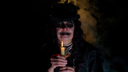 Sinister old elderly woman with horrible scary Halloween witch makeup in costume looking ominous at camera, making voodoo magic rituals with candle. Vampire granny isolated against black background