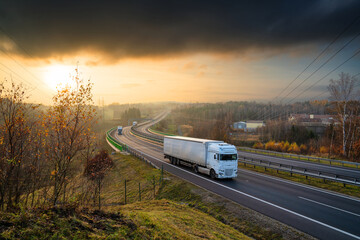 Three white truck driving on the highway winding through forested landscape in autumn colors at...