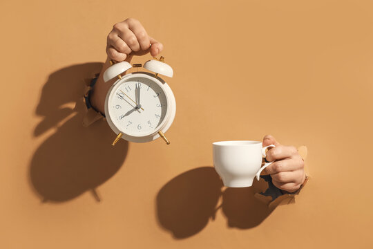 Female hands holding alarm clock and cup of coffee visible through hole on color background