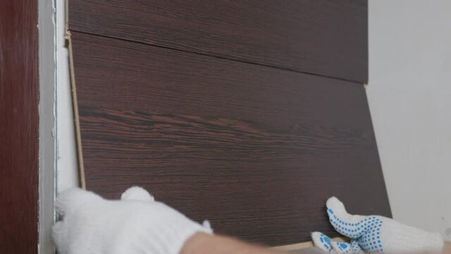 The specialist installs wall panels in the apartment. The brown panels are attached to wooden rails with glue. The repairman presses the panel hard for installation and leaves it.