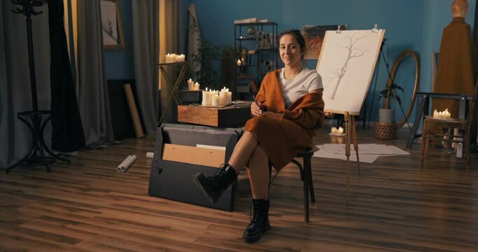 Woman with soul of an artist dressed in heavy black glans, long warm cardigan sits on chair in drawing studio, holding pencils in hand, in background is new sketch on easel depicting nature, a tree