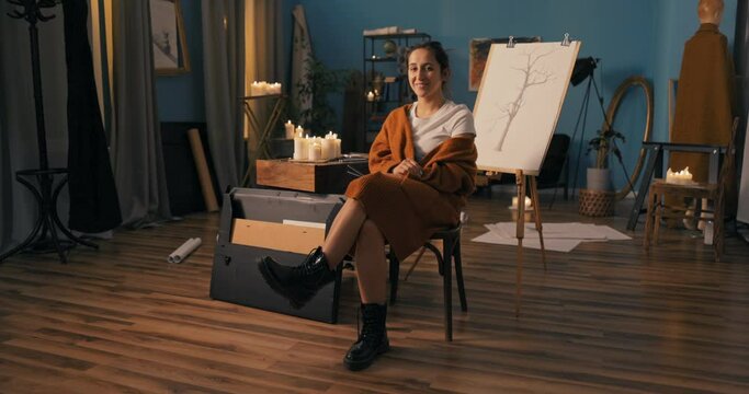 Woman with soul of an artist dressed in heavy black glans, long warm cardigan sits on chair in drawing studio, holding pencils in hand, in background is new sketch on easel depicting nature, a tree