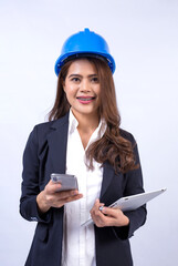 Beautiful Asian female engineer wearing black suit and blue safety helmet using smartphone and laptop computer isolated on gray in the studio background.