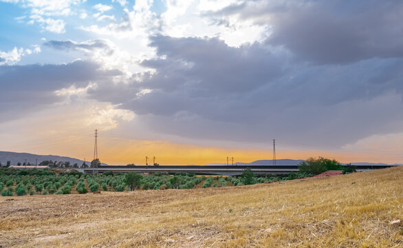 Las afueras de la ciudad al atardecer con trigo segado, olivos y una carretera y torres de electricidad contra un cielo naranja en el horizonte y azul por arriba separados por nubes oscuras de lluvia.