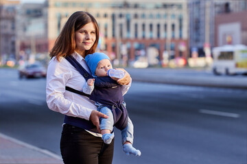 Mother is feeding baby in sling with bottle on street.