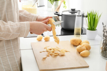 Woman peeling raw potatoes at table in kitchen, closeup