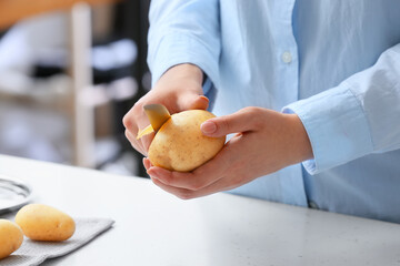 Woman peeling raw potatoes at table in kitchen, closeup