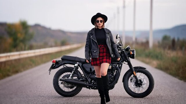 Sexy woman in red plaid skirt, leather jacket and hat walking to camera on vintage-styled motorcycle background. Attractive female motorcyclist in jackboots on highway. Trip, speed, freedom concept.