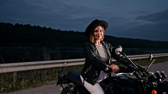 Hipster stylish motorcyclist woman sitting on vintage-styled motorcycle. Young female driver in hat at night on roadway. Trip, freedom, classic motorbike concept.