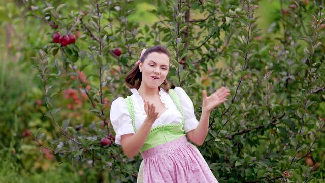 Comic woman in dirndl - traditional festival dress dancing funny and singing on ripe red apple autumn garden background. Dance, tomfoolery, positive mood and retro style concept.