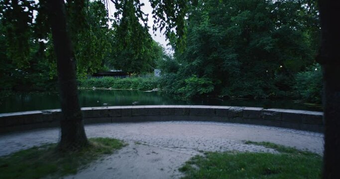 Tracking shot of a small circle shaped park with trees in the park (Friedrichsau) of Ulm. Camera tracks forward, revealing a small lake with ducks.