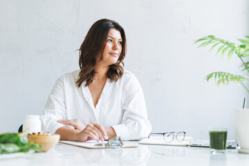Young smiling brunette woman doctor nutritionist plus size in white shirt working at laptop at modern bright office room. The doctor prescribes a prescription for medicines and vitamins at clinic