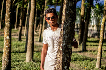 In the late afternoon sun, a boy peeks out from behind a tree and black sunglasses his eyes and behind the background blur.