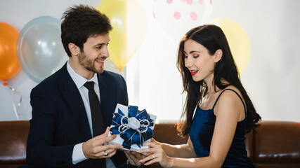 Caucasian bearded male businessman in formal business suit opening present gift box for happy beautiful female in casual dress in front colorful flying helium inflatable balloons in Birthday party