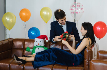 Obraz na płótnie Canvas Caucasian bearded businessman in formal business suit give present gift box to happy female girlfriend in long casual dress sitting on leather sofa with snowman doll with decoration colorful balloons