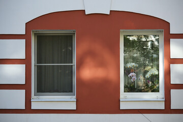Street facade of a residential building. Two windows with flowers in pots behind glass on a red and white painted wall.