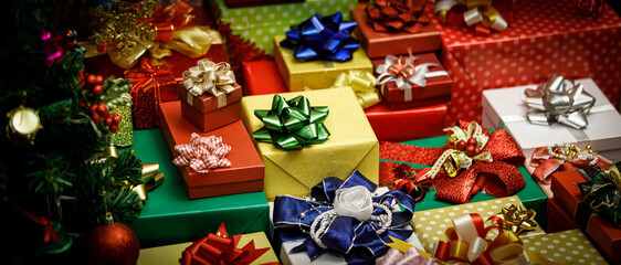 Close up shot of a lot of Merry Christmas eve wrapped present gift boxes stack with colorful ribbons and bows with decoration Xmas pine tree with glossy sphere balls bells socks ice flakes nearby © Bangkok Click Studio