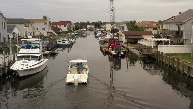 An aerial view of a fishing boat, slowly sailing through a canal behind houses on Long Island on a cloudy day. The camera dolly in behind the white boat as it moves forward past a crane on a barge.