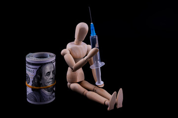 A wooden figurine of a conditional person with a syringe sits on a black background next to rolled-up 100USD bills