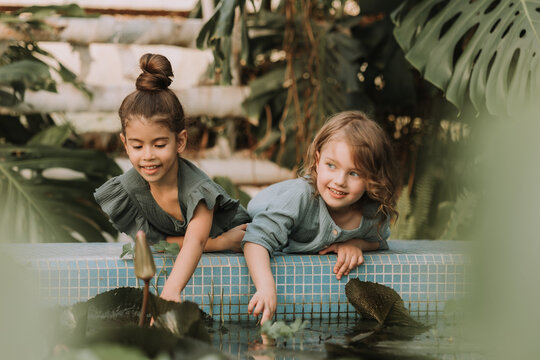 Two little girls are lying on the side of a pool with water plants in the greenhouse of the botanical garden surrounded by tropical leaves. High quality photo
