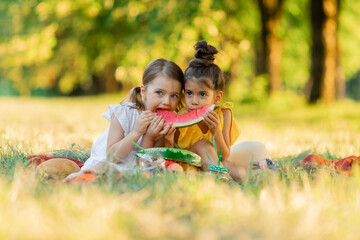 Girls are sitting on a blanket in the park and eat one slice of watermelon. Kids eat fruit outdoor. Healthy snack for children. Little girls are playing in the forest, biting a slice of watermelon.