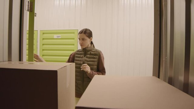 Locked-down view from inside storage container of young Caucasian woman opening door, putting in cardboard box and counting all boxes inside