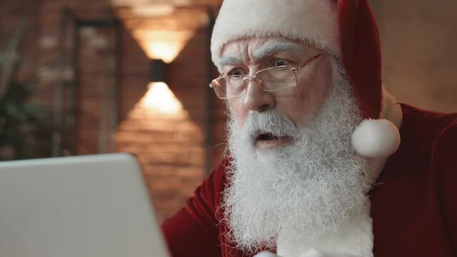 Close-up of white-bearded Santa Claus wearing eyeglasses, looking at cropped computer screen, getting shocked and surprised