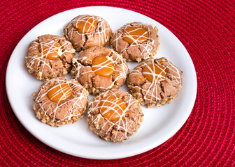Three brown cookies with hazelnuts, peanut brittle and lines of white cream on a white plate, caramel cream. White plate on red background