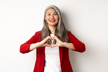 Beautiful asian mature woman in red blazer and makeup, showing heart sign and smiling, I love you...