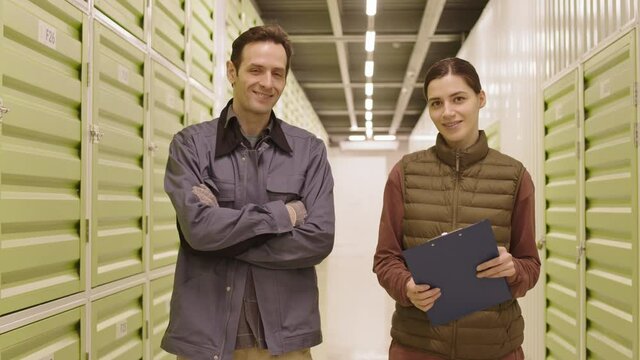 Medium of female and male Caucasian workers standing in hallway of storage facility, posing and smiling on camera, woman holding clipboard, man crossing arms on chest