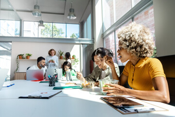 Two female coworkers have lunch while other colleagues work in the background. Copy space.