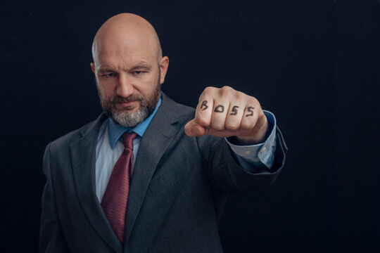 Portrait Of Bald Man In Business Suit On Dark Background. Gentleman In His 40s With Gray Beard. Boss Sign On His Fist.