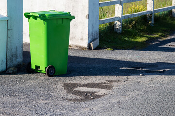 Naklejka premium Green plastic wheelie bin in a street ready for collection, waste and recycle industry. Warm sunny day