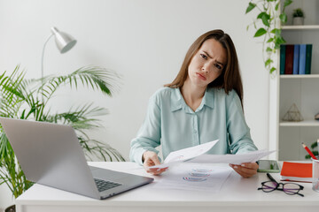 Bored sad young disappointed employee business woman in blue shirt hold paper account documents sit work at workplace white desk with laptop pc computer at office indoors. Achievement career concept